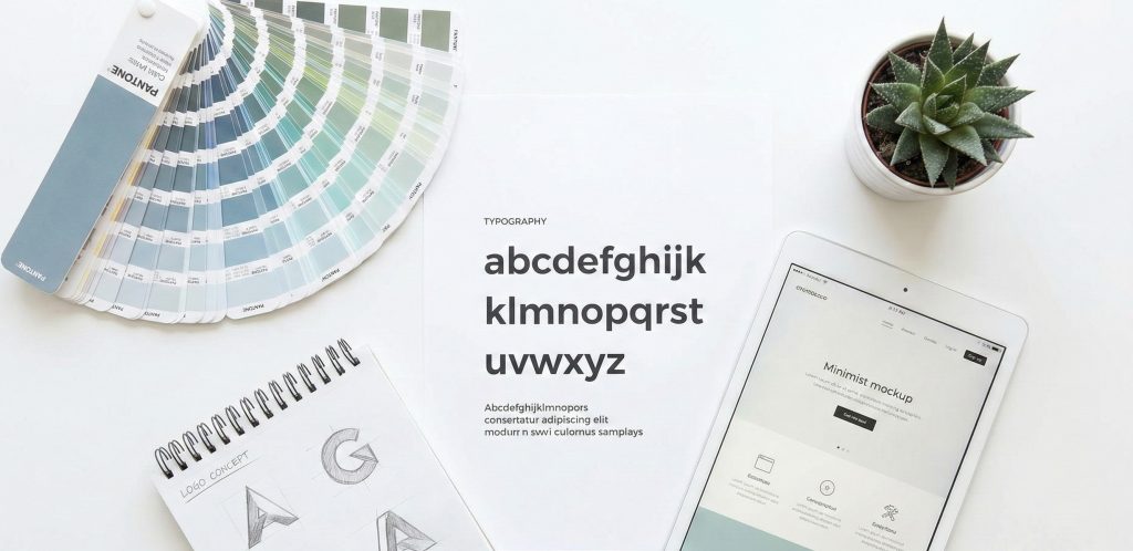 Ylhäältä otettu kuva suunnittelijan työpöydältä, jossa on esillä brändi-identiteetin osia: värikarttoja, fonttimalleja, logoluonnoksia lehtiössä sekä tabletti, jossa näkyy verkkosivuston malli. Kuva ilmentää yhtenäistä visuaalista suunnittelua.